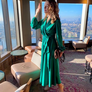 May include: A woman wearing a green wrap dress and gold heels stands in a modern lounge with a city view. She is holding a brown handbag.