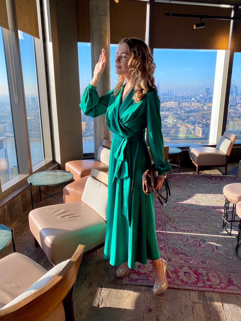May include: A woman wearing a green wrap dress and gold heels stands in a modern lounge with a city view. She is holding a brown handbag.