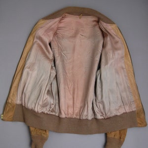 K&ouml;nnte beinhalten: Eine braune Wildlederbomberjacke mit Rei&szlig;verschluss und gerippten Strickb&uuml;ndchen und Bund. Die Jacke ist mit einem hellrosa Stoff gef&uuml;ttert.