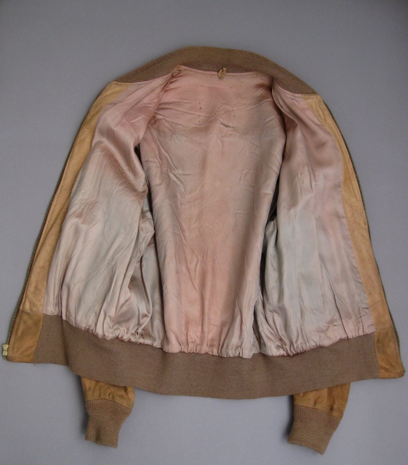 K&ouml;nnte beinhalten: Eine braune Wildlederbomberjacke mit Rei&szlig;verschluss und gerippten Strickb&uuml;ndchen und Bund. Die Jacke ist mit einem hellrosa Stoff gef&uuml;ttert.