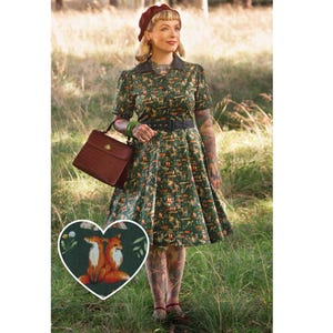 Op de afbeelding: Een vrouw draagt een groene jurk met een vos-print, een rode baret en een bruine leren handtas. De jurk heeft een getailleerd bovenstuk en een volle rok. De vrouw staat in een veld met groen gras.
