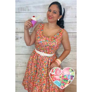 May include: A woman wearing a sleeveless dress with a colourful candy print. She is holding a milkshake with a cherry on top and a white belt around her waist.