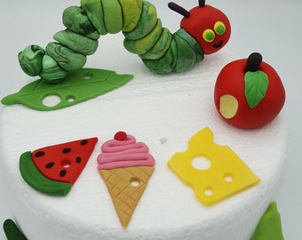 May include: A white cake decorated with a green caterpillar, a red apple, a slice of watermelon, a cone of ice cream, a slice of cheese, a pear, a strawberry, a cupcake, and a slice of orange. The cake is decorated with fondant.