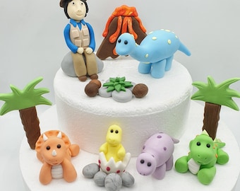 May include: A cake decorated with colorful dinosaur-themed fondant figures. The scene includes a volcano, palm trees, a seated explorer, and various dinosaurs in shades of orange, blue, green, and purple. The cake is on a white tiered base.