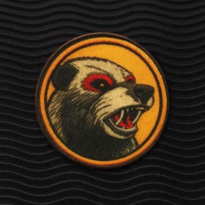 Könnte beinhalten: Runder Aufnäher mit einem knurrenden Bären mit roten Augen und freiliegenden Zähnen. Der Bär ist vor einem gelben Hintergrund mit braunem Rand platziert. Der Aufnäher befindet sich auf einem schwarzen, strukturierten Hintergrund.