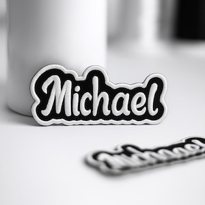 May include: A black and white embroidered patch featuring the name Michael in a cursive script font. The patch has a thick black background with a white border and white lettering, with a second identical patch blurred in the foreground.