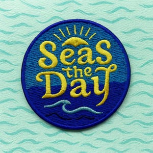 Puede incluir: Un parche redondo bordado con la frase "Seas the Day" en escritura amarilla. El parche presenta un fondo azul con un sol amarillo y un diseño de olas. El fondo es azul claro con un patrón de olas.