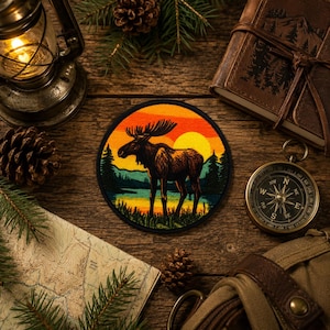 May include: A circular embroidered patch featuring a dark brown moose standing in a grassy field near a lake, set against a vibrant orange and yellow sunset sky with silhouettes of pine trees, framed by a thick black border.
