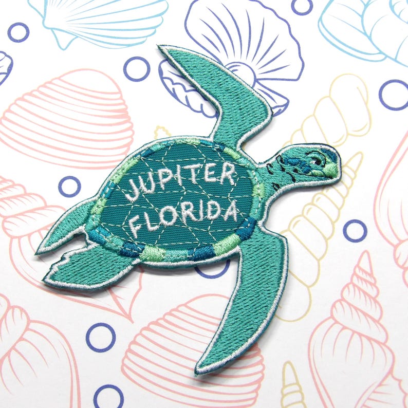 Pu&ograve; includere: Toppa ricamata a forma di tartaruga marina turchese con la scritta "Jupiter Florida". La toppa ha un bordo bianco ed &egrave; ambientata su uno sfondo di conchiglie e cerchi blu.