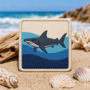 May include: Square patch featuring a dark gray shark swimming above blue waves. The patch has a beige border and background. The image is set on a sandy beach with seashells, driftwood, and a blurred ocean in the background.