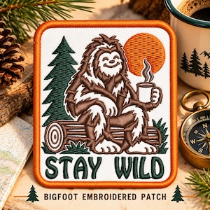 May include: A rectangular embroidered patch with an orange border featuring a brown Bigfoot sitting on a log, holding a steaming mug. A dark green pine tree and an orange sun are in the background. The text STAY WILD is in dark green.