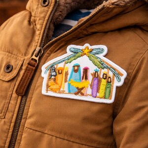 May include: A rectangular embroidered patch featuring a colourful nativity scene with a yellow star at the top, attached to a brown jacket. The design includes stylised figures in blue, purple, yellow, and green, all set under a green roof structure.