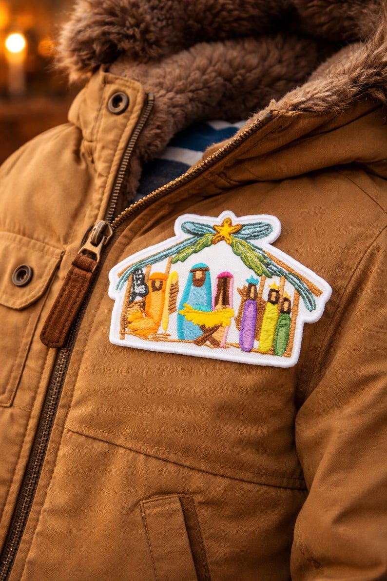 May include: A rectangular embroidered patch featuring a colourful nativity scene with a yellow star at the top, attached to a brown jacket. The design includes stylised figures in blue, purple, yellow, and green, all set under a green roof structure.