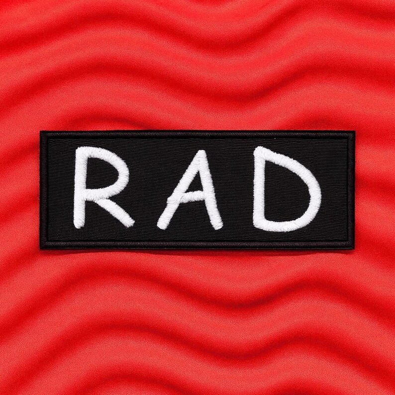 May include: A rectangular black embroidered patch featuring the word RAD in white capital letters. The patch has a thick black border and is centred on a vibrant red background with a wavy, textured surface pattern.