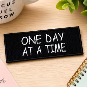 Puede incluir: Un parche rectangular negro bordado con el texto blanco ONE DAY AT A TIME en una fuente sans-serif sencilla. El parche tiene un borde cosido limpio y se presenta como un artículo plano y horizontal con un mensaje claro y legible.