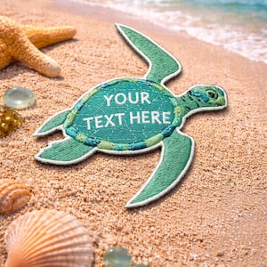 Puede incluir: Un parche de tortuga bordada verde con el texto "YOUR TEXT HERE" en una playa de arena con conchas marinas, una estrella de mar y piedras de vidrio transparente. El océano es visible en el fondo.