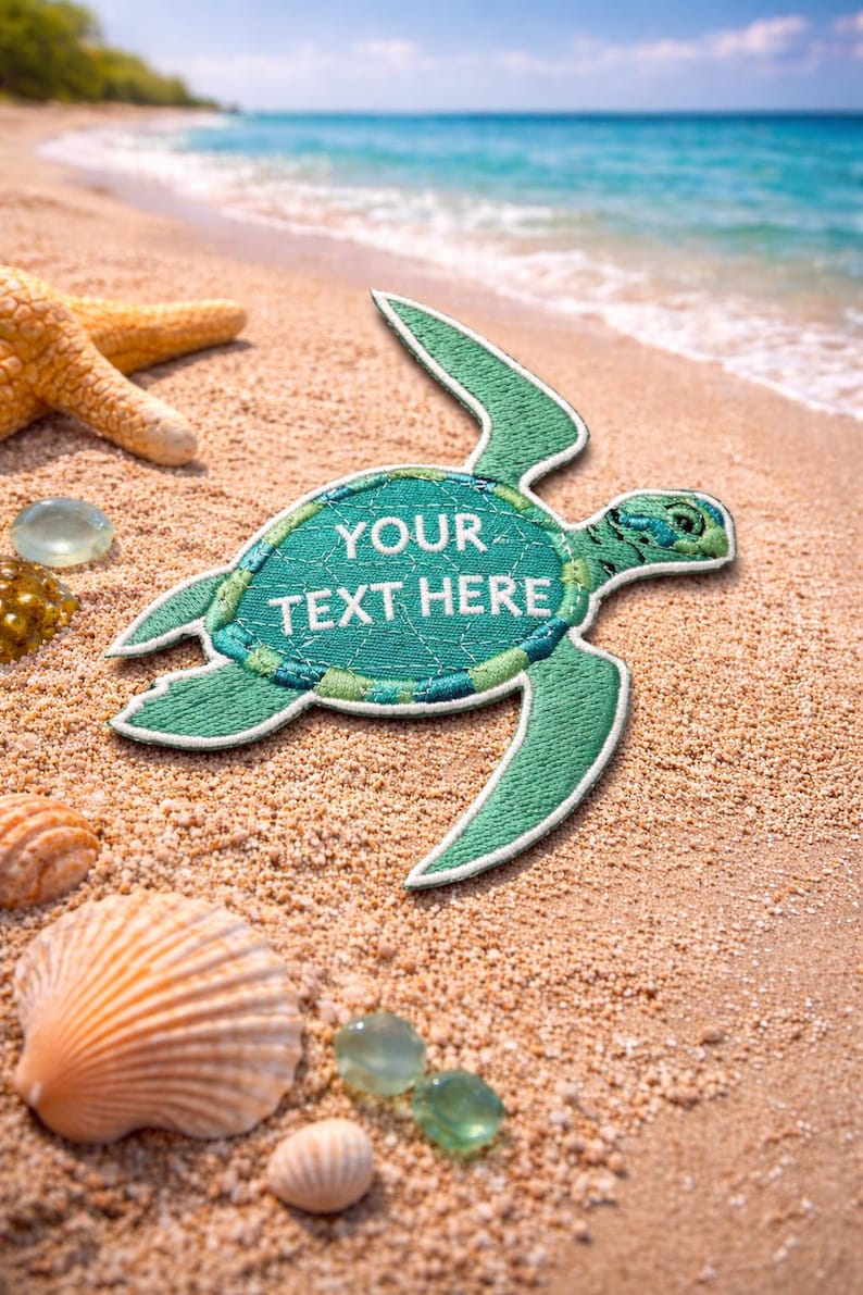 Pu&ograve; includere: Una toppa a forma di tartaruga ricamata verde con la scritta "YOUR TEXT HERE" su una spiaggia sabbiosa con conchiglie, una stella marina e pietre di vetro trasparente. L'oceano &egrave; visibile sullo sfondo.