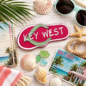 Op de afbeelding: Een roze geborduurde patch in de vorm van een slipper met een groene band en de witte tekst KEY WEST. De patch is omringd door schelpen, een ansichtkaart van Key West, een zonnebril, een spiraalblok en een touw op een witte ondergrond.