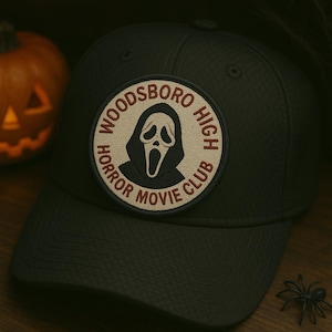 Może przedstawiać: Ciemnoszara czapka z daszkiem z okrągłą naszywką z napisem "Woodsboro High Horror Movie Club" wokół białej twarzy ducha. W tle wyrzeźbiona dynia i plastikowy pająk.