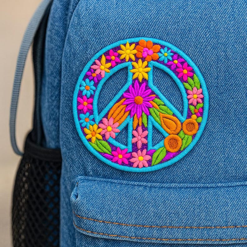 K&ouml;nnte beinhalten: Ein blauer Jeans-Rucksack mit einem farbenfrohen, gestickten Peace-Zeichen-Patch. Der Patch hat einen t&uuml;rkisfarbenen Umriss mit einem floralen Design in Rosa-, Orange-, Gelb- und Gr&uuml;nt&ouml;nen. Der Rucksack hat eine schwarze Netztasche.