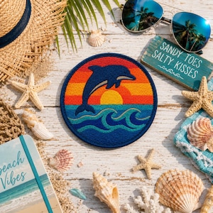 May include: A circular embroidered patch featuring a dark blue dolphin leaping over stylized ocean waves against a sunset background of orange, yellow, and red horizontal stripes, with a dark blue border surrounding the entire circular design.