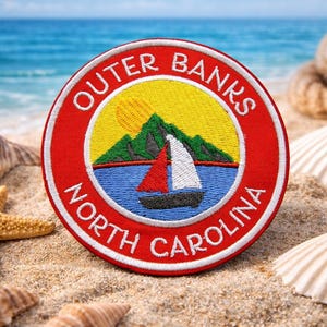 May include: Embroidered patch with a red border and the words "Outer Banks North Carolina" in white. The patch features a sailboat on blue water, a yellow sun, and green mountains. Seashells and a starfish are in the sand.