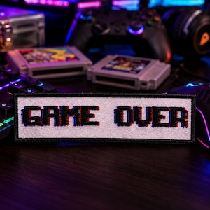May include: A rectangular embroidered patch featuring the text GAME OVER in a pixelated black font with red and blue shadow accents on a white background. The patch has a black border and is positioned horizontally in the center of the frame.