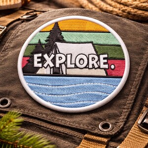 May include: A circular embroidered patch with a camping theme. The patch features a mountain scene with a tent, trees, and the word "EXPLORE." The design includes blue, green, yellow, and white colors, with a white border. The patch is on a brown fabric.