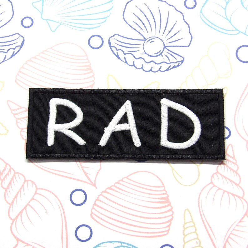 May include: A rectangular black embroidered patch featuring the word RAD in bold white capital letters. The patch has a clean, stitched border and is presented against a background pattern of light-coloured seashells and small circles.