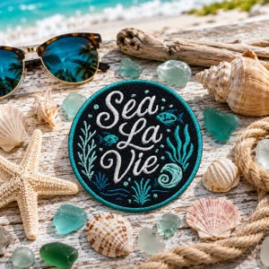 May include: A circular embroidered patch featuring the white cursive text Sea La Vie surrounded by teal aquatic illustrations of fish, seaweed, and shells on a black background, accented with a teal border and a detailed stitched finish.