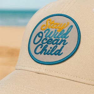 Op de afbeelding: Beige baseballpet met een ronde patch. De patch heeft de tekst "Stay Wild Ocean Child" in geel en blauw, omgeven door een blauwe rand. De pet staat tegen een vage strandachtergrond.