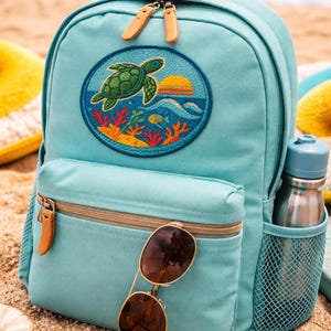 May include: A light blue backpack with a turtle patch, a front pocket, and a side mesh pocket. The backpack has a tan handle and zipper pulls. Sunglasses rest on the front pocket. A water bottle is in the side pocket.