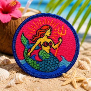 May include: A circular embroidered patch depicting a mermaid with red hair, a yellow torso, and a green tail, holding a gold trident. The mermaid is set against a pink background with a blue border. The patch is on a sandy beach with seashells.