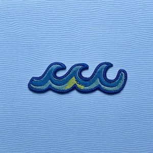 May include: Embroidered wave patch in shades of blue with yellow accents. The patch has a dark blue outline, giving a raised effect. It's set against a light blue, textured background. The design evokes the ocean and coastal themes.