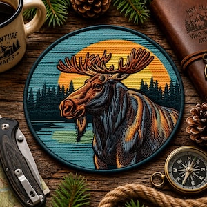 May include: A circular embroidered patch featuring a moose in profile against a yellow sun and dark forest background. The moose has large antlers and a textured coat in shades of brown, blue, and orange. The patch has a dark blue border.