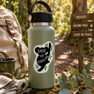 May include: A sage green water bottle with a black handle and lid. A sticker on the bottle reads "Save the Koalas" with a koala silhouette and green leaves. The bottle is set in a natural outdoor scene with a backpack and trail sign.