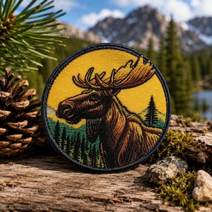 May include: A circular embroidered patch featuring a brown moose in profile against a vibrant yellow sky. The lower portion of the patch depicts a green forest landscape with a single tall pine tree. The entire design is bordered by a black stitched edge.