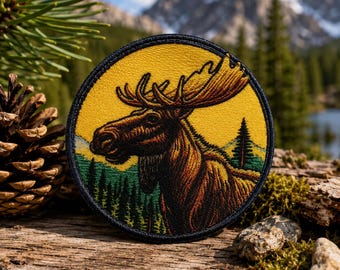May include: A circular embroidered patch featuring a brown moose in profile against a vibrant yellow sky. The lower portion of the patch depicts a green forest landscape with a single tall pine tree. The entire design is bordered by a black stitched edge.
