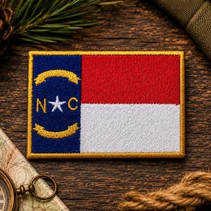 May include: A rectangular embroidered patch featuring the North Carolina state flag. The design includes a blue vertical bar on the left with a white star between the letters N and C, flanked by two gold scrolls, and red and white horizontal stripes.