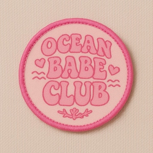 Puede incluir: Un parche redondo rosa con las palabras "OCEAN BABE CLUB" en letras de burbujas. El parche tiene un borde rosa y presenta corazones, olas y un diseño floral. El fondo es de tela rosa claro.