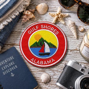 Peut inclure: Un écusson brodé circulaire avec une bordure rouge portant le texte blanc GULF SHORES ALABAMA. Le centre représente un soleil jaune, des montagnes vertes et un voilier aux voiles rouge et blanche sur une eau bleue, entouré de coquillages.