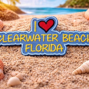 Może przedstawiać: Niebieska naszywka z żółtym napisem "I Clearwater Beach Florida" i czerwonym sercem. Naszywka otoczona jest muszlami i piaskiem, z plażą i oceanem w tle.