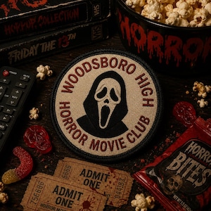 May include: A circular embroidered patch featuring a beige background with a black hooded ghost mask graphic in the center. The text "WOODSBORO HIGH HORROR MOVIE CLUB" is stitched in dark red around the border of the patch on a beige fabric.