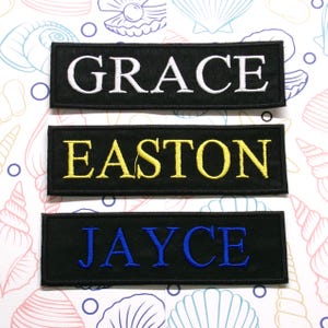 Puede incluir: Tres parches de tela negra con letras bordadas en blanco, amarillo y azul. Los parches dicen "GRACE", "EASTON" y "JAYCE".