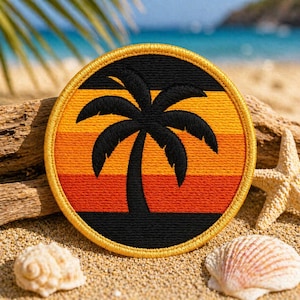 Puede incluir: Parche bordado redondo con una silueta de palmera negra sobre un degradado de atardecer naranja, amarillo y negro. El parche tiene un borde dorado. El parche está sobre arena con conchas marinas y madera flotante.