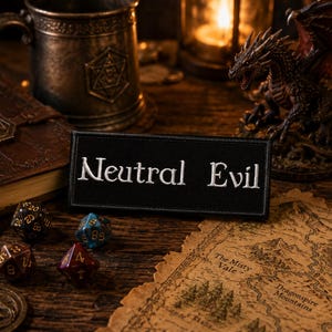 Peut inclure: Un écusson rectangulaire en tissu noir avec le texte Neutral Evil brodé en blanc. L'écusson présente une bordure cousue nette et est positionné au centre, affichant un contraste clair entre le fond sombre et les lettres blanches.