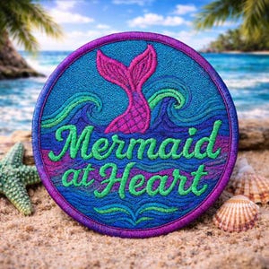 Puede incluir: Parche bordado con temática de sirena. El parche redondo presenta un borde morado, fondo azul y una cola de sirena rosa. Las palabras "Mermaid at Heart" están en verde. El parche está en una playa de arena con conchas marinas.
