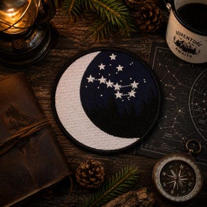 May include: A circular embroidered patch featuring a white crescent moon against a dark navy blue night sky. The design includes a cluster of white stitched stars forming a constellation above a silhouette of dark pine trees at the bottom edge.