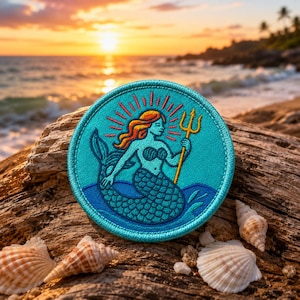 May include: A circular turquoise patch depicting a mermaid with red hair and a golden trident. The mermaid is set against a blue sea design. The patch is on a piece of driftwood, with seashells scattered around it.