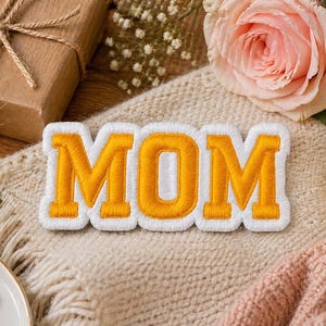 Puede incluir: Un parche bordado blanco y dorado con la palabra "MOM" en letras de bloque en negrita. El parche está sobre una superficie de punto color crema, con una rosa rosa, una vela encendida y un regalo envuelto en el fondo. El parche es un artículo decorativo.